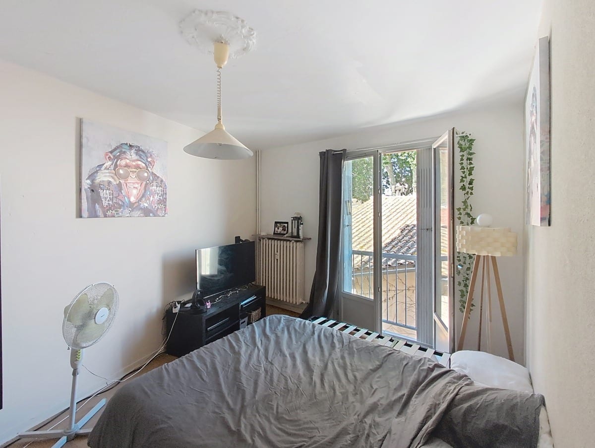 Chambre avec lit double recouvert d'un drap gris, meuble TV noir, ventilateur sur pied et lampadaire tripode en bois. Porte-fenêtre avec balcon donnant sur vue des toits, rosace décorative au plafond et tableaux colorés aux murs.