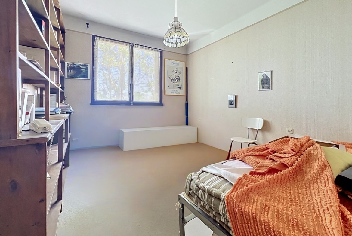Chambre avec lit simple recouvert d'un couvre-lit orange côtelé et étagères en bois foncé le long du mur gauche. Fenêtre avec encadrement bleu, sol clair et suspension style Tiffany au plafond. Affiches encadrées dont une de Picasso au mur.