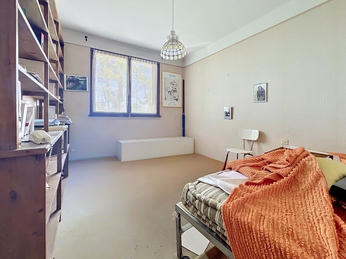 Chambre avec lit simple recouvert d'un couvre-lit orange côtelé et étagères en bois foncé le long du mur gauche. Fenêtre avec encadrement bleu, sol clair et suspension style Tiffany au plafond. Affiches encadrées dont une de Picasso au mur.