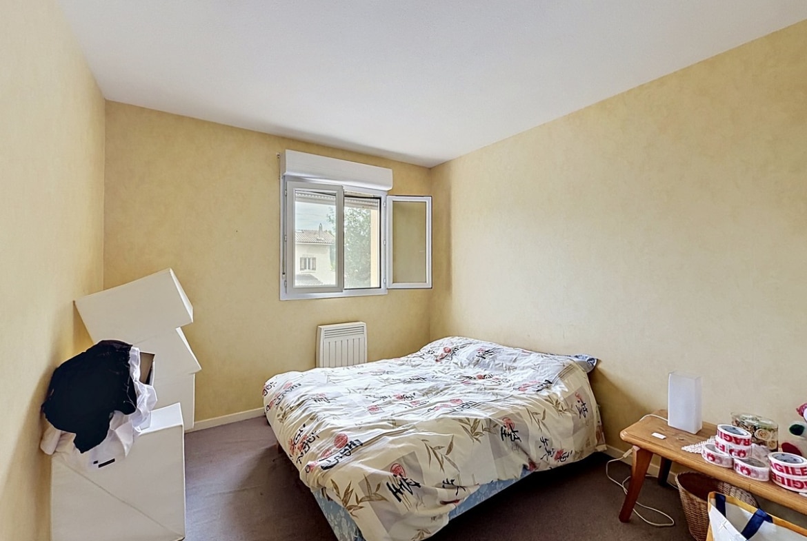 Chambre avec murs enduits jaune ocre et moquette marron au sol. Lit double avec couette à motifs floraux, fenêtre à deux vantaux avec volet roulant blanc, mobilier de déménagement visible incluant cartons et petit meuble en bois.