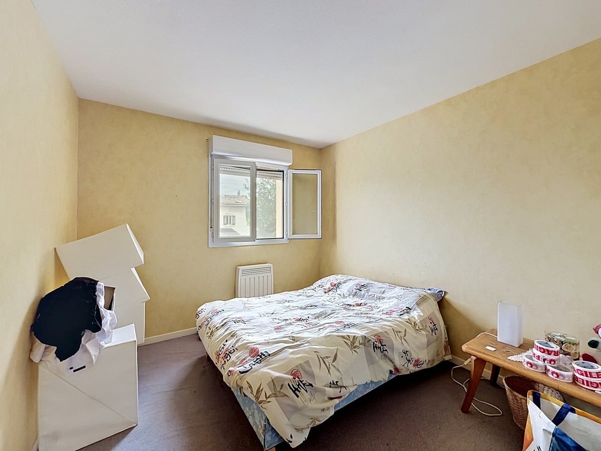 Chambre avec murs enduits jaune ocre et moquette marron au sol. Lit double avec couette à motifs floraux, fenêtre à deux vantaux avec volet roulant blanc, mobilier de déménagement visible incluant cartons et petit meuble en bois.