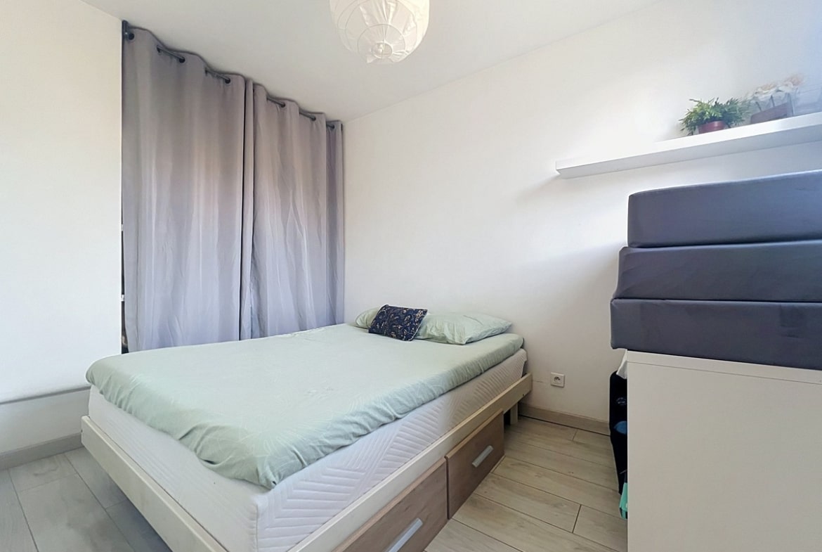 Chambre avec lit double en bois clair équipé de tiroirs de rangement, linge de lit vert d'eau. Rideaux gris occultants, étagère murale blanche et sol en parquet stratifié clair.
