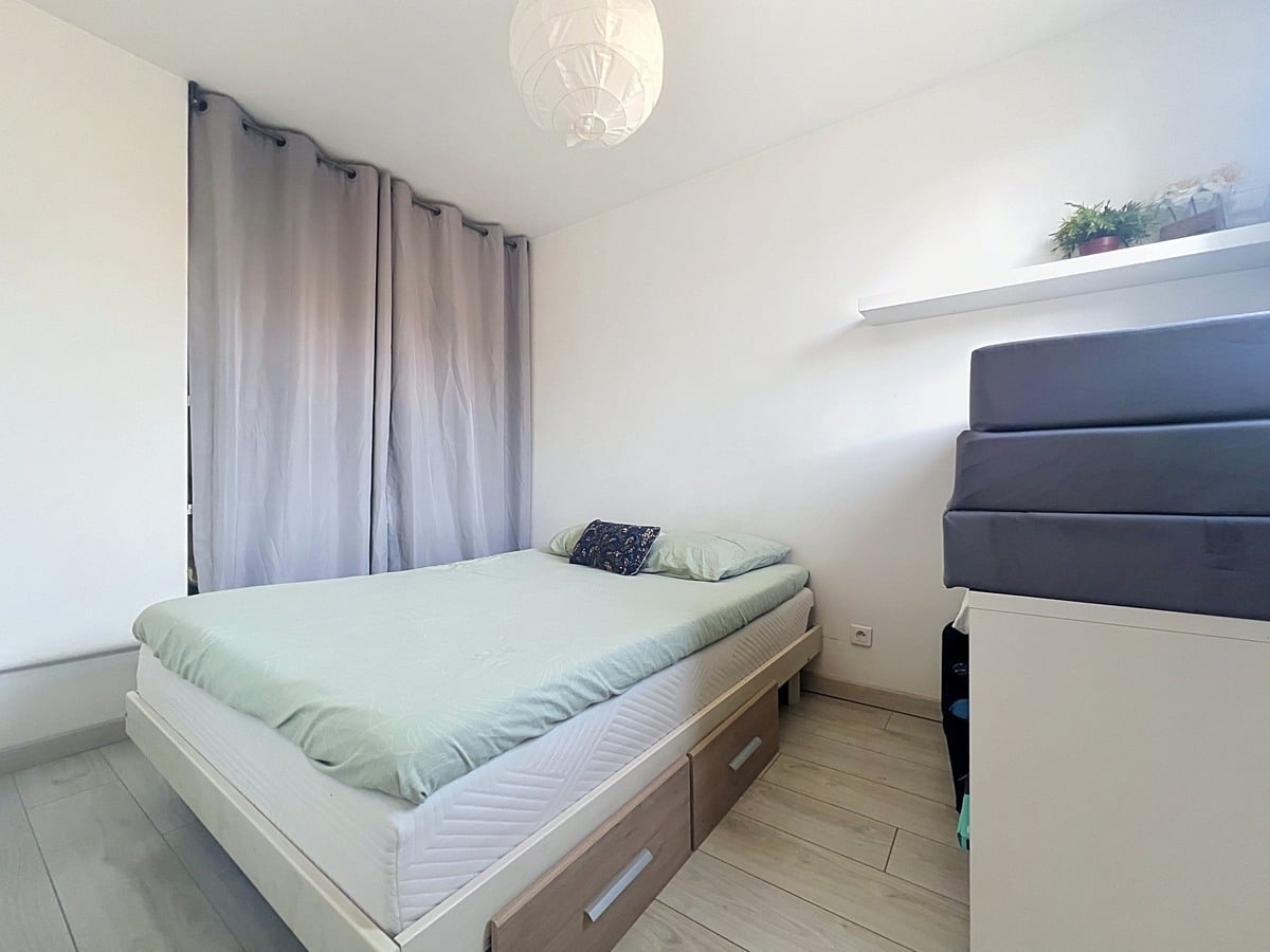 Chambre avec lit double en bois clair équipé de tiroirs de rangement, linge de lit vert d'eau. Rideaux gris occultants, étagère murale blanche et sol en parquet stratifié clair.