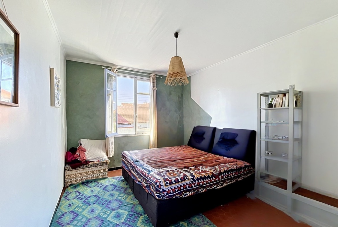 Chambre avec lit double sur cadre bleu foncé et couverture à motifs géométriques. Mur d'accent vert olive, tapis bleu-vert à motifs ethniques, étagère blanche avec livres et fenêtre à double battant donnant sur les toits en tuiles.