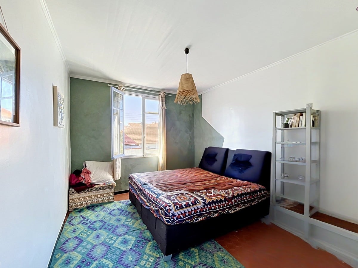 Chambre avec lit double sur cadre bleu foncé et couverture à motifs géométriques. Mur d'accent vert olive, tapis bleu-vert à motifs ethniques, étagère blanche avec livres et fenêtre à double battant donnant sur les toits en tuiles.