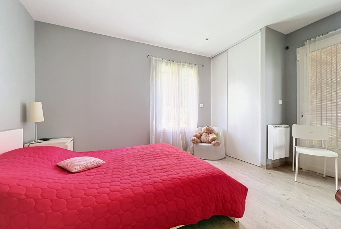 Chambre avec murs gris et parquet clair, lit double recouvert d'une couette rouge vif. Placard intégré avec portes coulissantes blanches, fauteuil blanc avec peluche, bureau avec store vénitien et radiateur électrique mural.