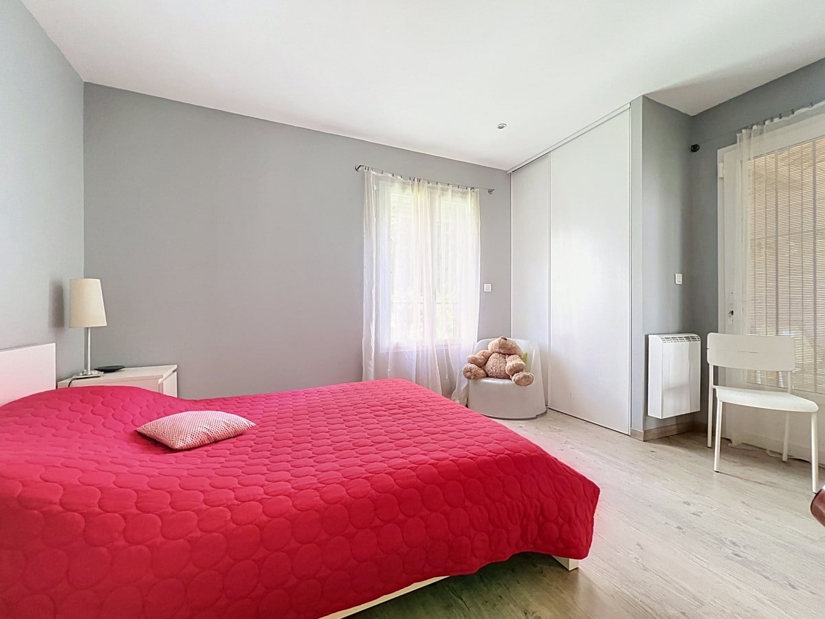 Chambre avec murs gris et parquet clair, lit double recouvert d'une couette rouge vif. Placard intégré avec portes coulissantes blanches, fauteuil blanc avec peluche, bureau avec store vénitien et radiateur électrique mural.