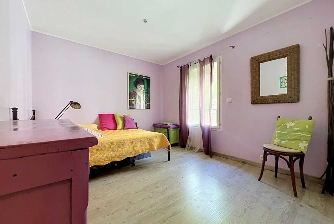 Chambre avec murs violet pâle et parquet clair, lit simple avec couvre-lit jaune et coussins colorés. Bureau en bois peint bordeaux, chaise bistrot avec coussin vert, miroir en osier tressé et affiche du film Amélie Poulain au mur.
