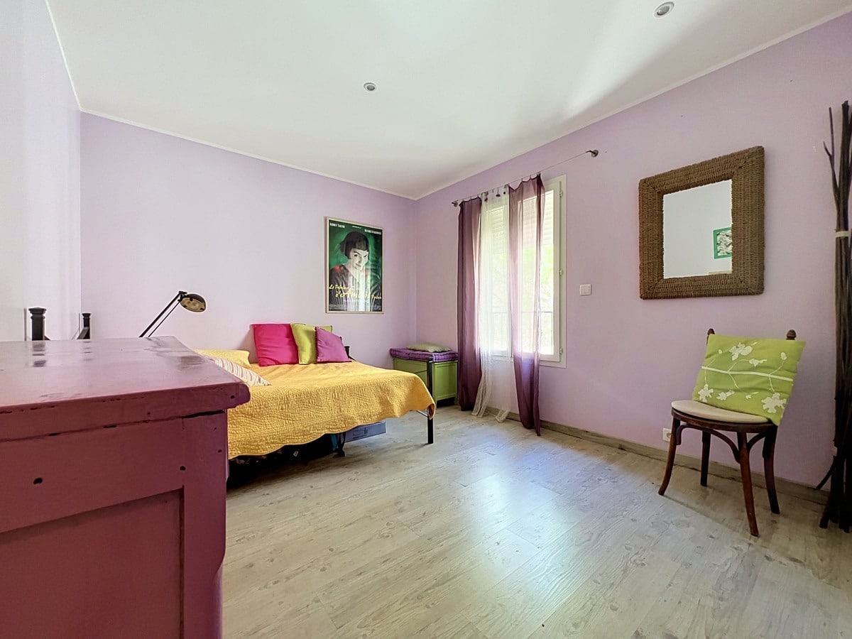 Chambre avec murs violet pâle et parquet clair, lit simple avec couvre-lit jaune et coussins colorés. Bureau en bois peint bordeaux, chaise bistrot avec coussin vert, miroir en osier tressé et affiche du film Amélie Poulain au mur.