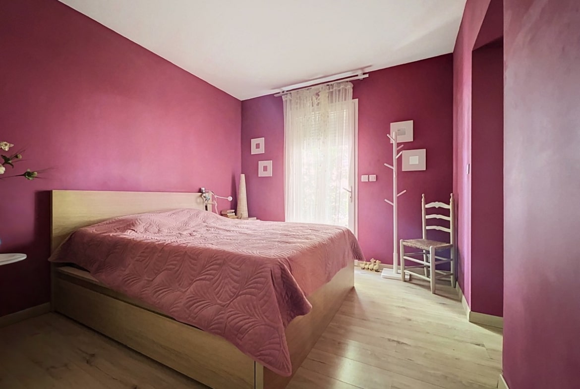 Chambre avec murs peints en rose fuchsia et parquet clair en bois. Lit double avec cadre en bois naturel, couvre-lit rose pâle à motifs de feuilles, chaise traditionnelle blanche et porte-manteau blanc en forme d'arbre.
