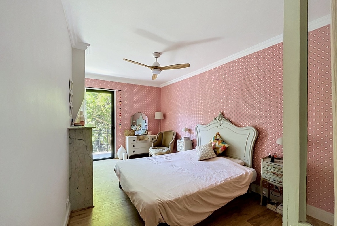Chambre avec mur en papier peint rose à motifs géométriques et tête de lit sculptée blanc crème de style classique. Parquet bois clair, cheminée en marbre gris, ventilateur de plafond à pales bois et porte-fenêtre donnant sur verdure extérieure.