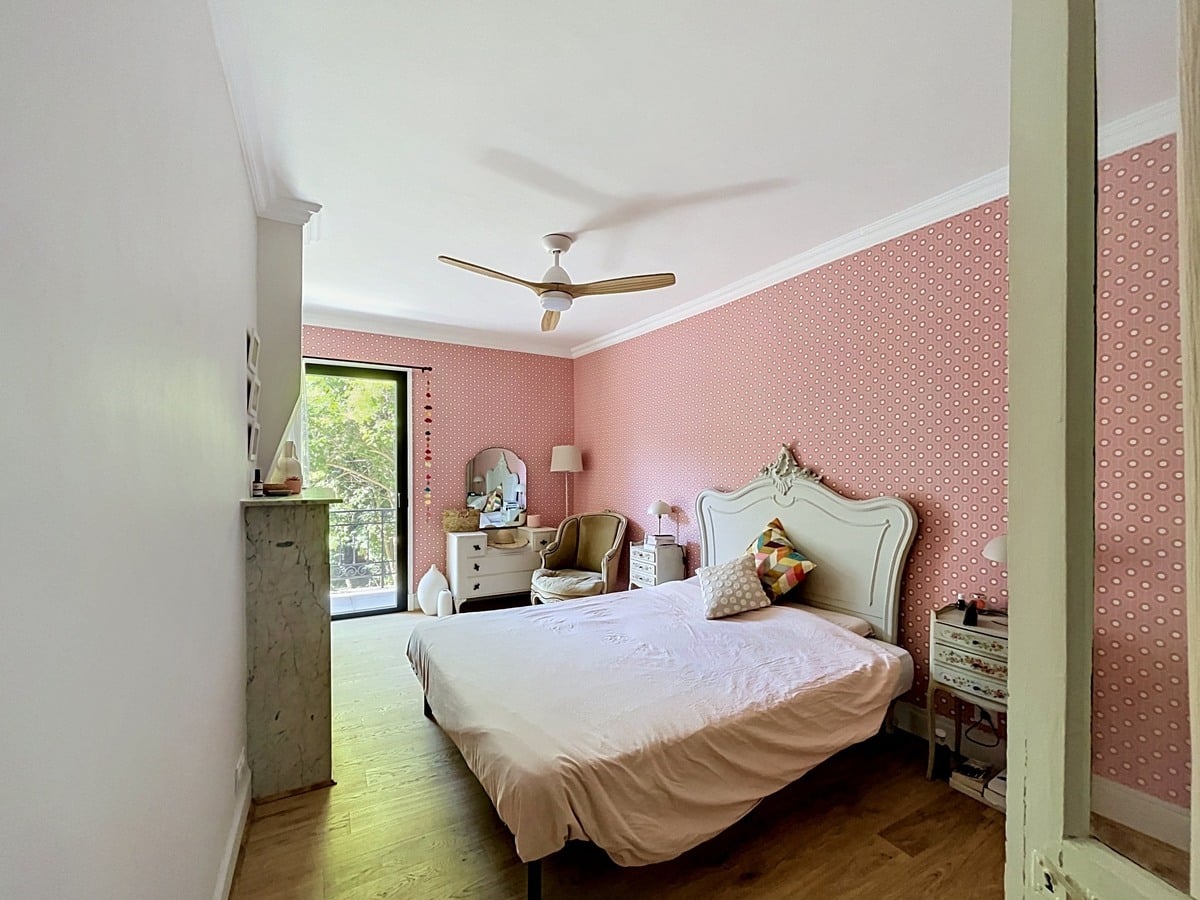 Chambre avec mur en papier peint rose à motifs géométriques et tête de lit sculptée blanc crème de style classique. Parquet bois clair, cheminée en marbre gris, ventilateur de plafond à pales bois et porte-fenêtre donnant sur verdure extérieure.