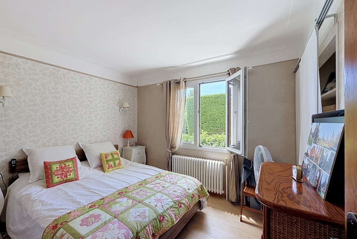 Chambre avec lit double et couette à motifs floraux verts et roses. Sol en parquet clair, radiateur en fonte blanc sous la fenêtre ouverte, papier peint bicolore beige et gris.