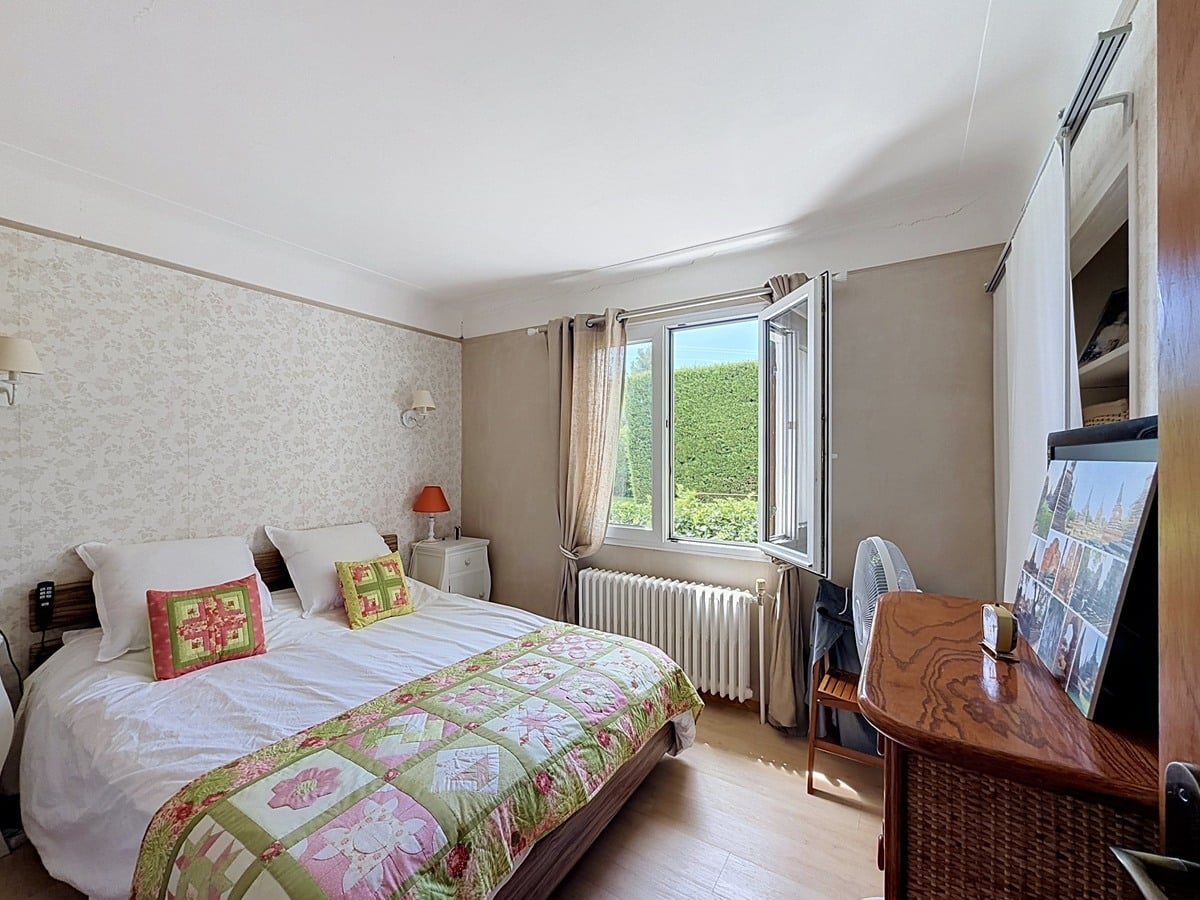 Chambre avec lit double et couette à motifs floraux verts et roses. Sol en parquet clair, radiateur en fonte blanc sous la fenêtre ouverte, papier peint bicolore beige et gris.