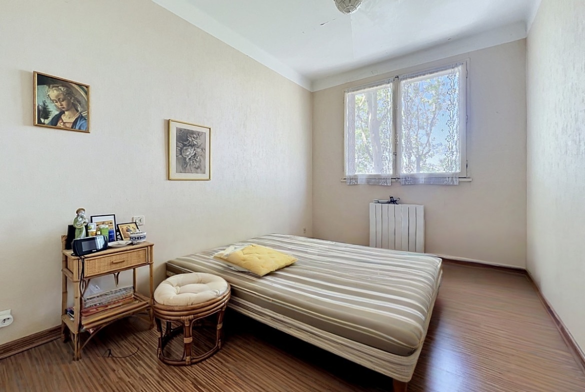 Chambre avec lit double à matelas rayé beige et parquet stratifié brun. Table de chevet en rotin, petit pouf assorti et radiateur blanc sous la fenêtre. Murs en papier peint crème texturé avec deux cadres anciens accrochés.