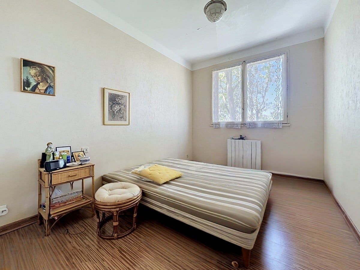 Chambre avec lit double à matelas rayé beige et parquet stratifié brun. Table de chevet en rotin, petit pouf assorti et radiateur blanc sous la fenêtre. Murs en papier peint crème texturé avec deux cadres anciens accrochés.