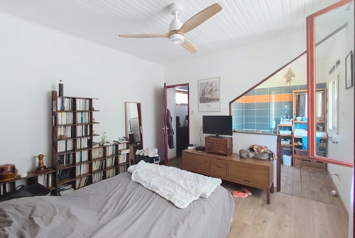 Chambre avec lit double couverture grise, parquet clair et plafond en lambris blanc avec ventilateur. Vue sur salle d'eau attenante avec carrelage bleu et orange, meuble TV en bois et bibliothèque murale.