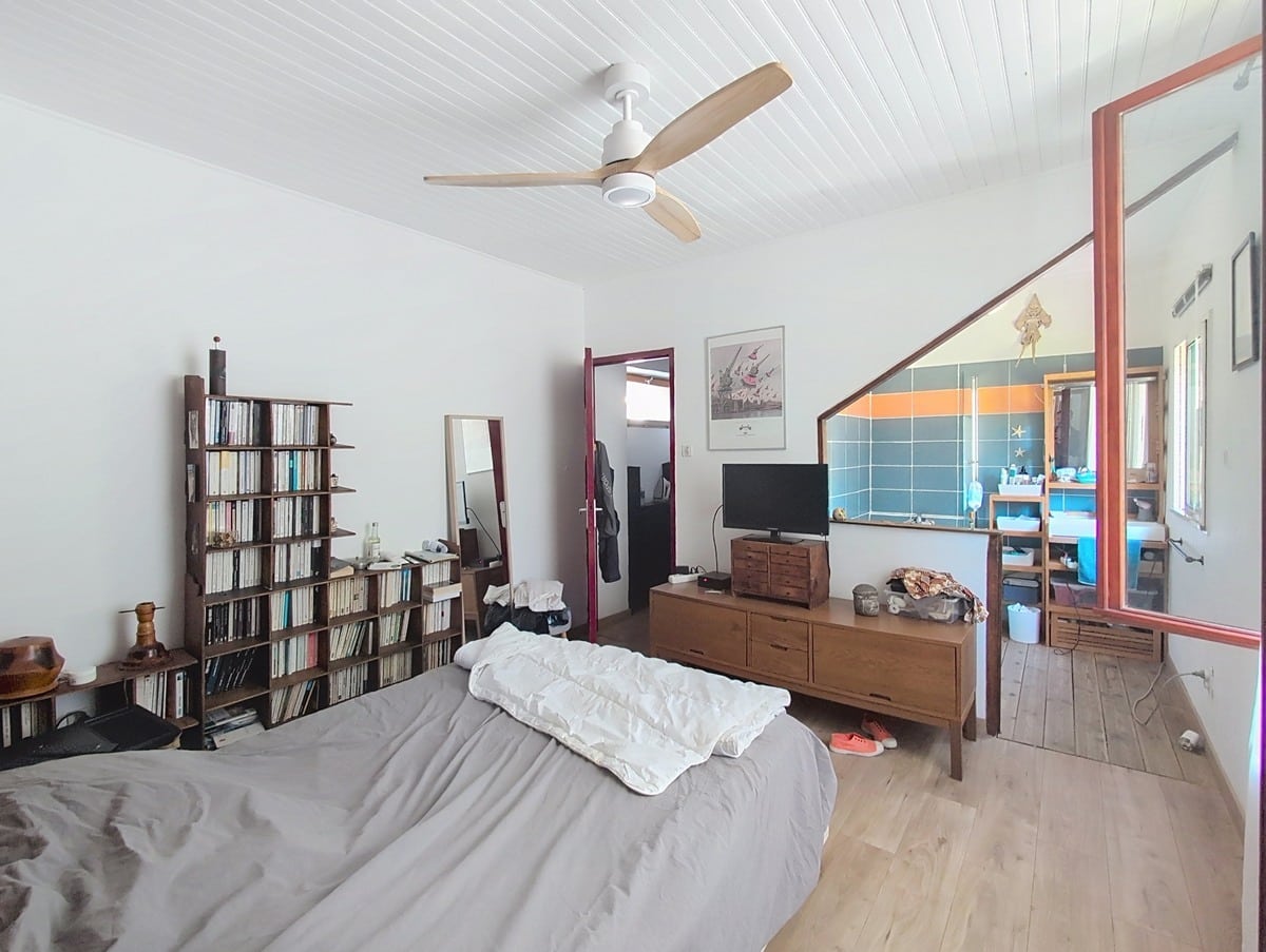 Chambre avec lit double couverture grise, parquet clair et plafond en lambris blanc avec ventilateur. Vue sur salle d'eau attenante avec carrelage bleu et orange, meuble TV en bois et bibliothèque murale.