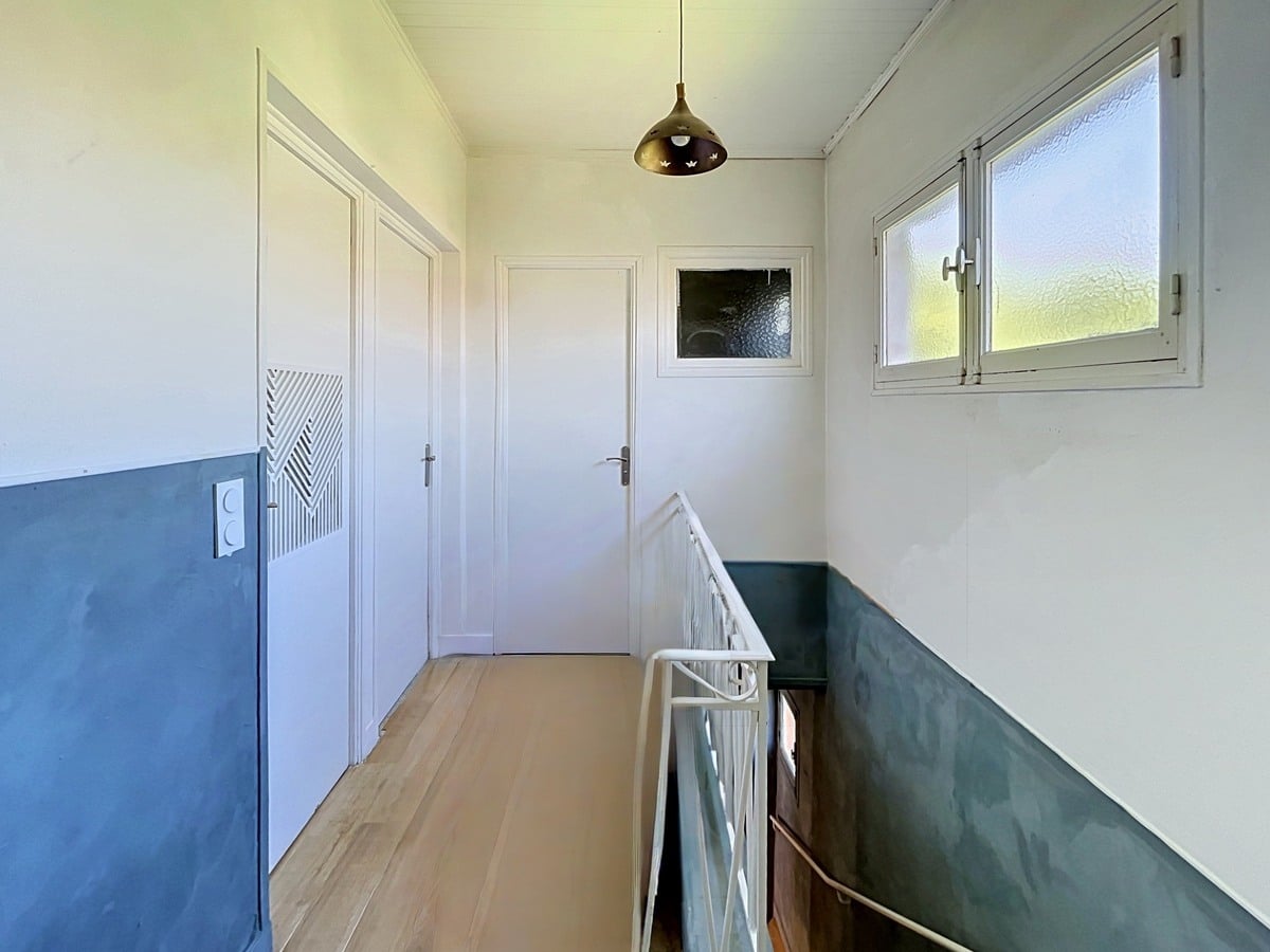 Couloir d'étage avec parquet clair et soubassement bleu sur un mur, vert sur l'autre. Garde-corps métallique blanc menant à l'escalier, trois portes blanches et fenêtres en verre dépoli diffusant la lumière naturelle.
