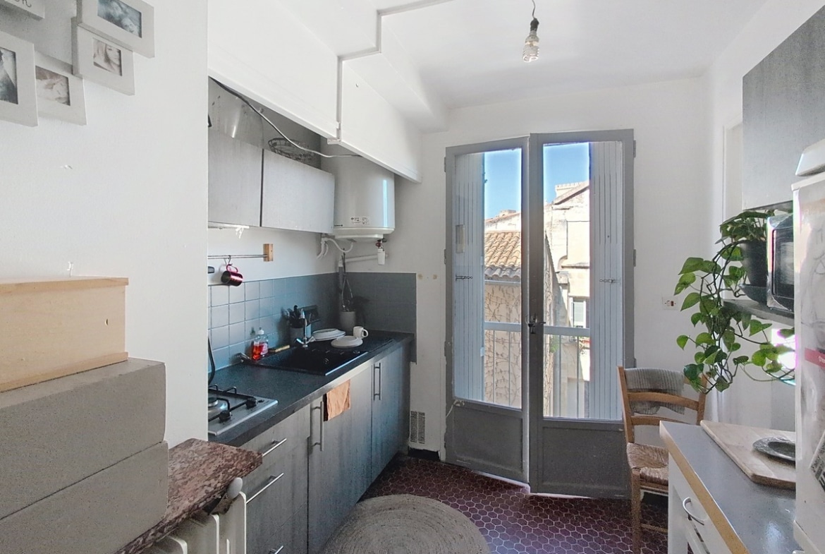 Cuisine avec meubles gris anthracite, crédence carrelée bleue, plaque de cuisson gaz et chauffe-eau mural. Sol en tomettes hexagonales bordeaux, porte-fenêtre à petits carreaux ouverte sur vue des toits en tuiles provençales.