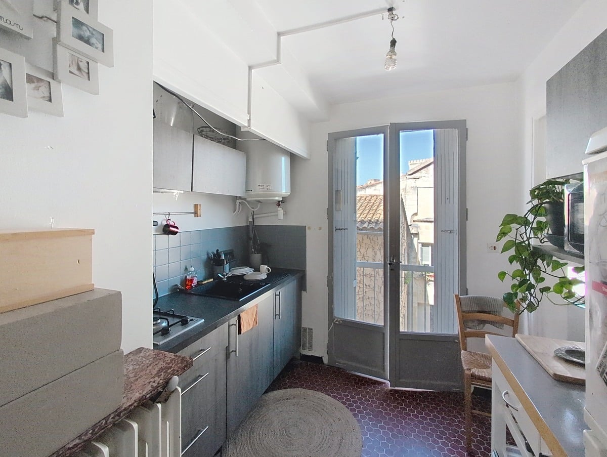 Cuisine avec meubles gris anthracite, crédence carrelée bleue, plaque de cuisson gaz et chauffe-eau mural. Sol en tomettes hexagonales bordeaux, porte-fenêtre à petits carreaux ouverte sur vue des toits en tuiles provençales.