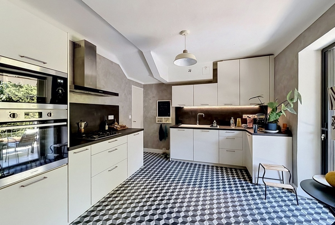 Cuisine moderne en L avec façades blanches mates, plan de travail noir et crédence en béton ciré gris. Sol en carreaux de ciment à motifs géométriques noir blanc gris, four et micro-ondes encastrés, plaque gaz et fenêtre sur verdure.