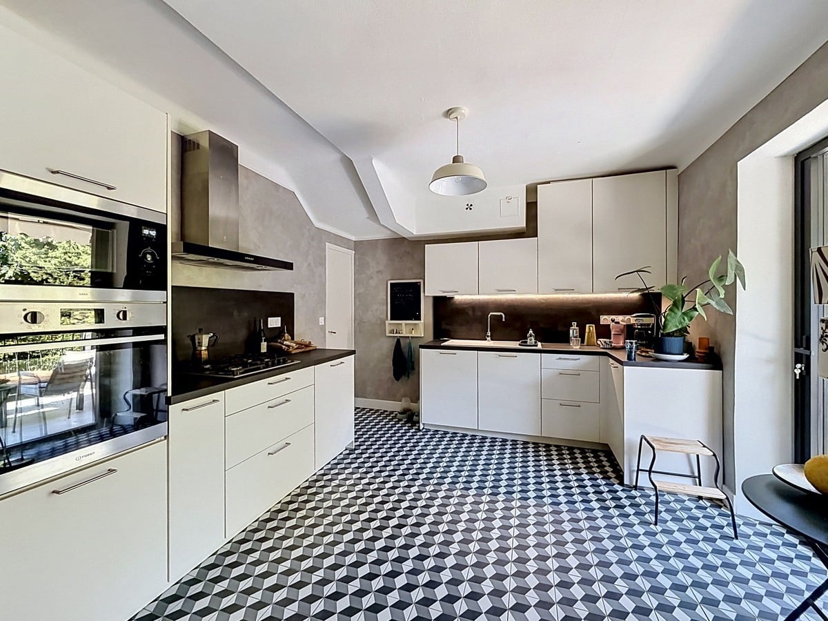 Cuisine moderne en L avec façades blanches mates, plan de travail noir et crédence en béton ciré gris. Sol en carreaux de ciment à motifs géométriques noir blanc gris, four et micro-ondes encastrés, plaque gaz et fenêtre sur verdure.