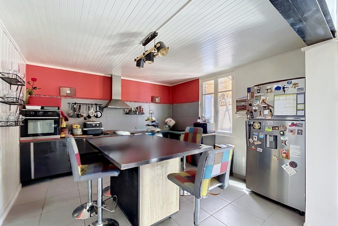Cuisine aménagée avec murs rouges, meubles bas noirs et plan de travail sombre. Îlot central en bois clair avec tabourets multicolores, réfrigérateur américain inox et plafond en lambris blanc avec spots orientables.