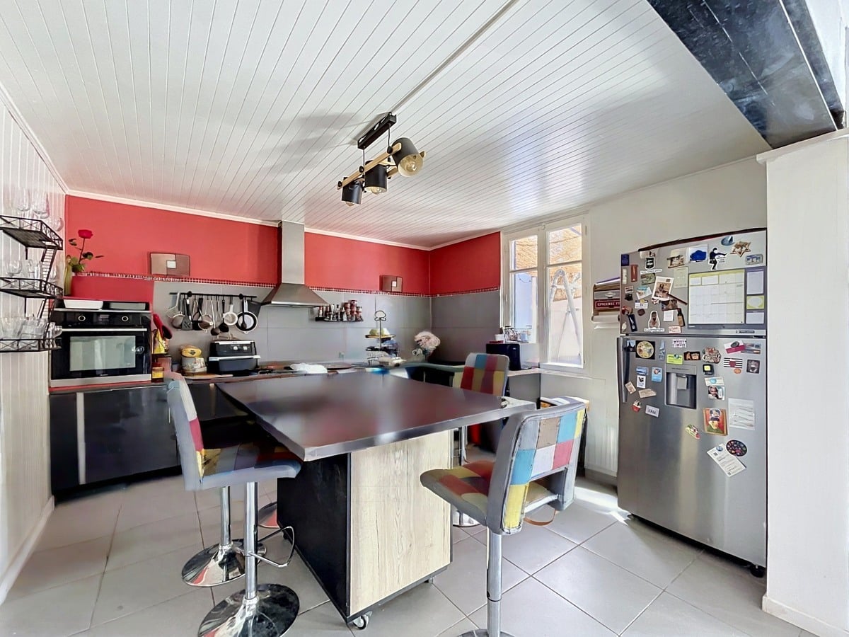 Cuisine aménagée avec murs rouges, meubles bas noirs et plan de travail sombre. Îlot central en bois clair avec tabourets multicolores, réfrigérateur américain inox et plafond en lambris blanc avec spots orientables.
