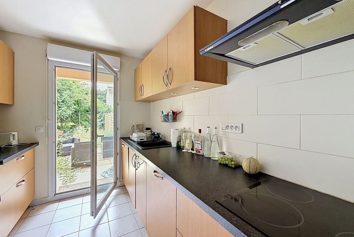 Cuisine en longueur avec meubles hauts et bas en bois clair et plan de travail en granit noir. Équipements intégrés comprenant plaque vitrocéramique et hotte aspirante, carrelage blanc au mur et porte-fenêtre ouvrant sur le jardin.