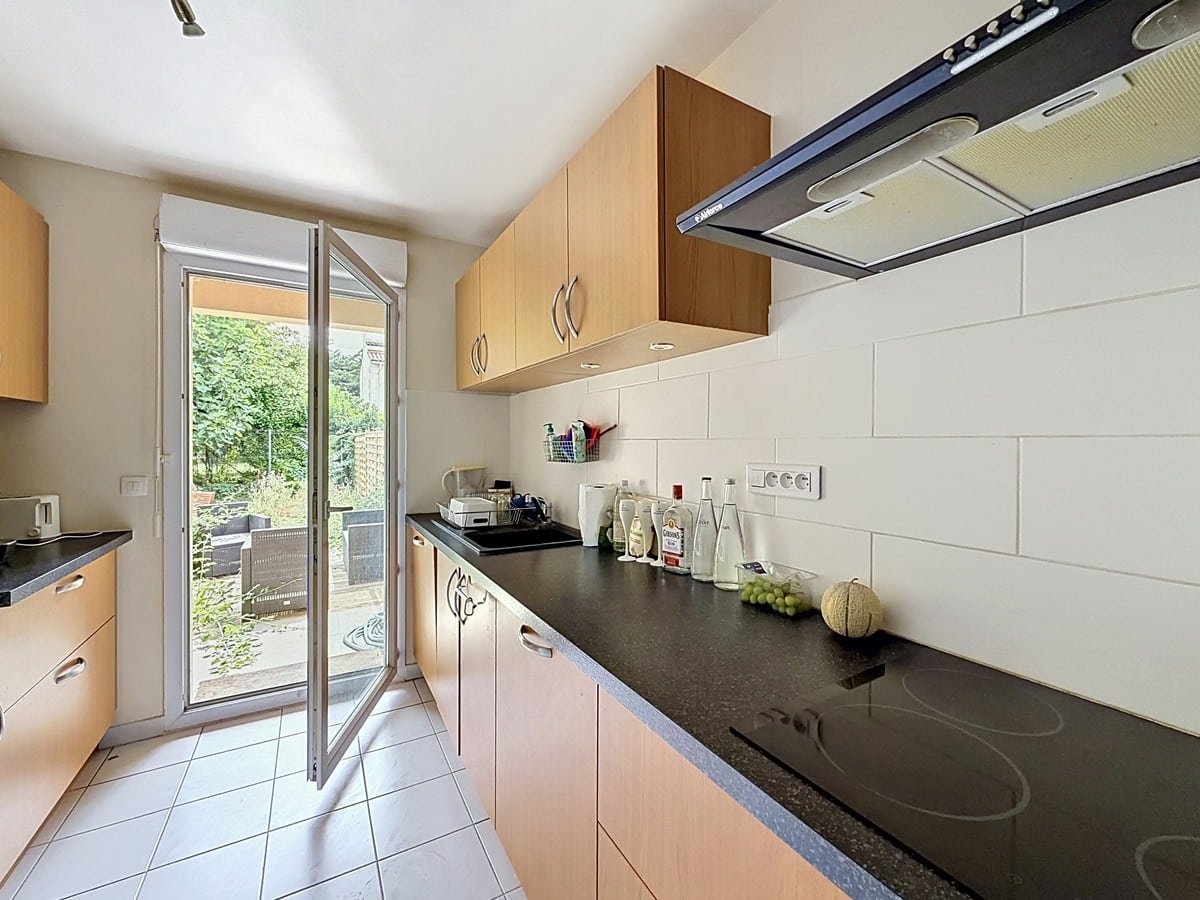 Cuisine en longueur avec meubles hauts et bas en bois clair et plan de travail en granit noir. Équipements intégrés comprenant plaque vitrocéramique et hotte aspirante, carrelage blanc au mur et porte-fenêtre ouvrant sur le jardin.