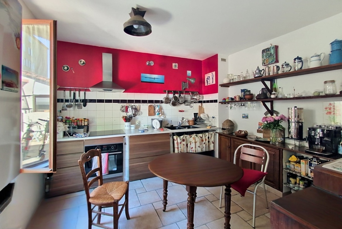 Cuisine aménagée avec meubles en bois foncé, mur d'accent rouge vif et crédence carrelée blanche. Table ronde en bois avec chaises paillées, étagères ouvertes garnies de vaisselle et accessoires de cuisine.