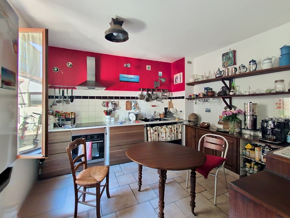 Cuisine aménagée avec meubles en bois foncé, mur d'accent rouge vif et crédence carrelée blanche. Table ronde en bois avec chaises paillées, étagères ouvertes garnies de vaisselle et accessoires de cuisine.