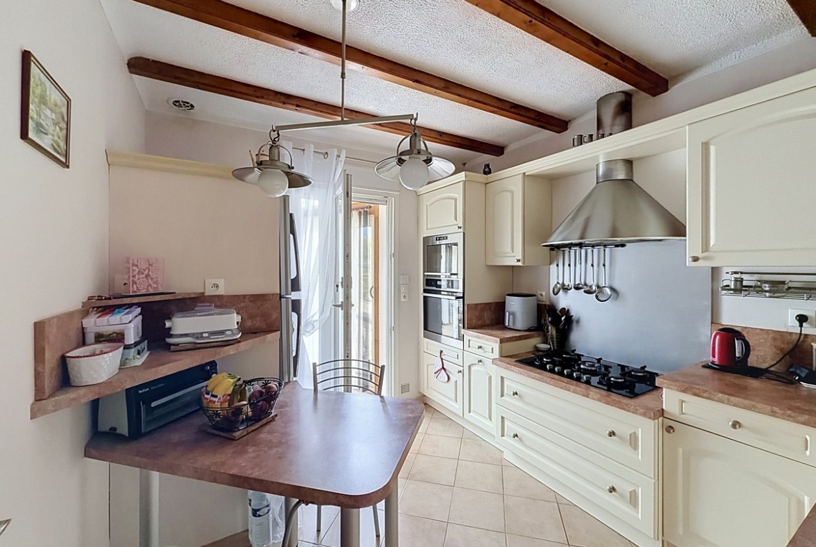 Cuisine aménagée avec meubles en bois laqué crème et plan de travail marron. Plaque gaz, hotte inox, four encastré et réfrigérateur américain. Poutres en bois au plafond et carrelage beige au sol.