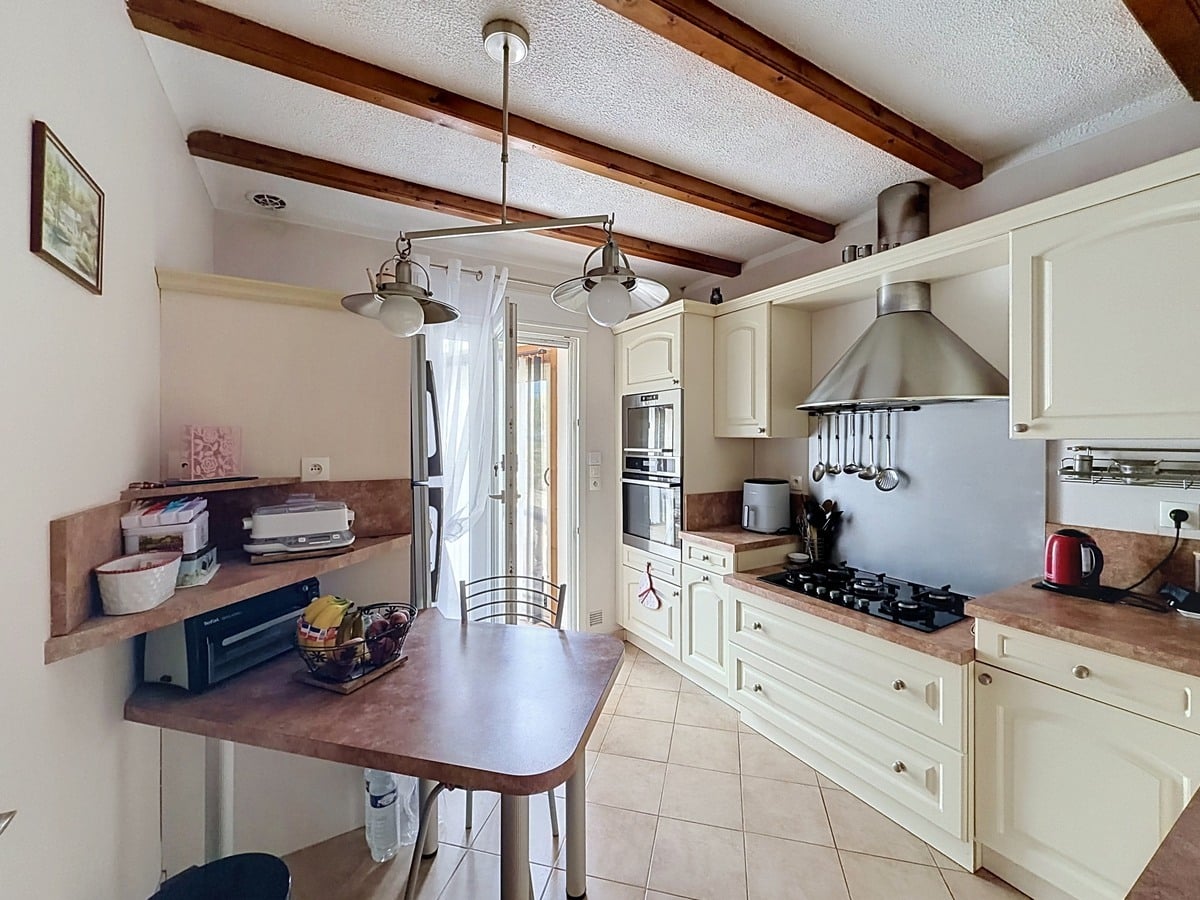Cuisine aménagée avec meubles en bois laqué crème et plan de travail marron. Plaque gaz, hotte inox, four encastré et réfrigérateur américain. Poutres en bois au plafond et carrelage beige au sol.