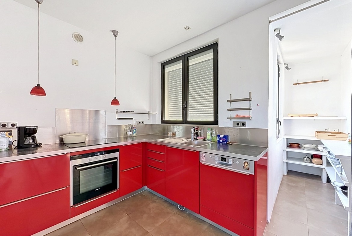 Cuisine en L avec meubles rouge brillant, plan de travail et crédence en inox brossé. Four encastré, lave-vaisselle intégré, suspensions rouges et cellier attenant visible à droite.