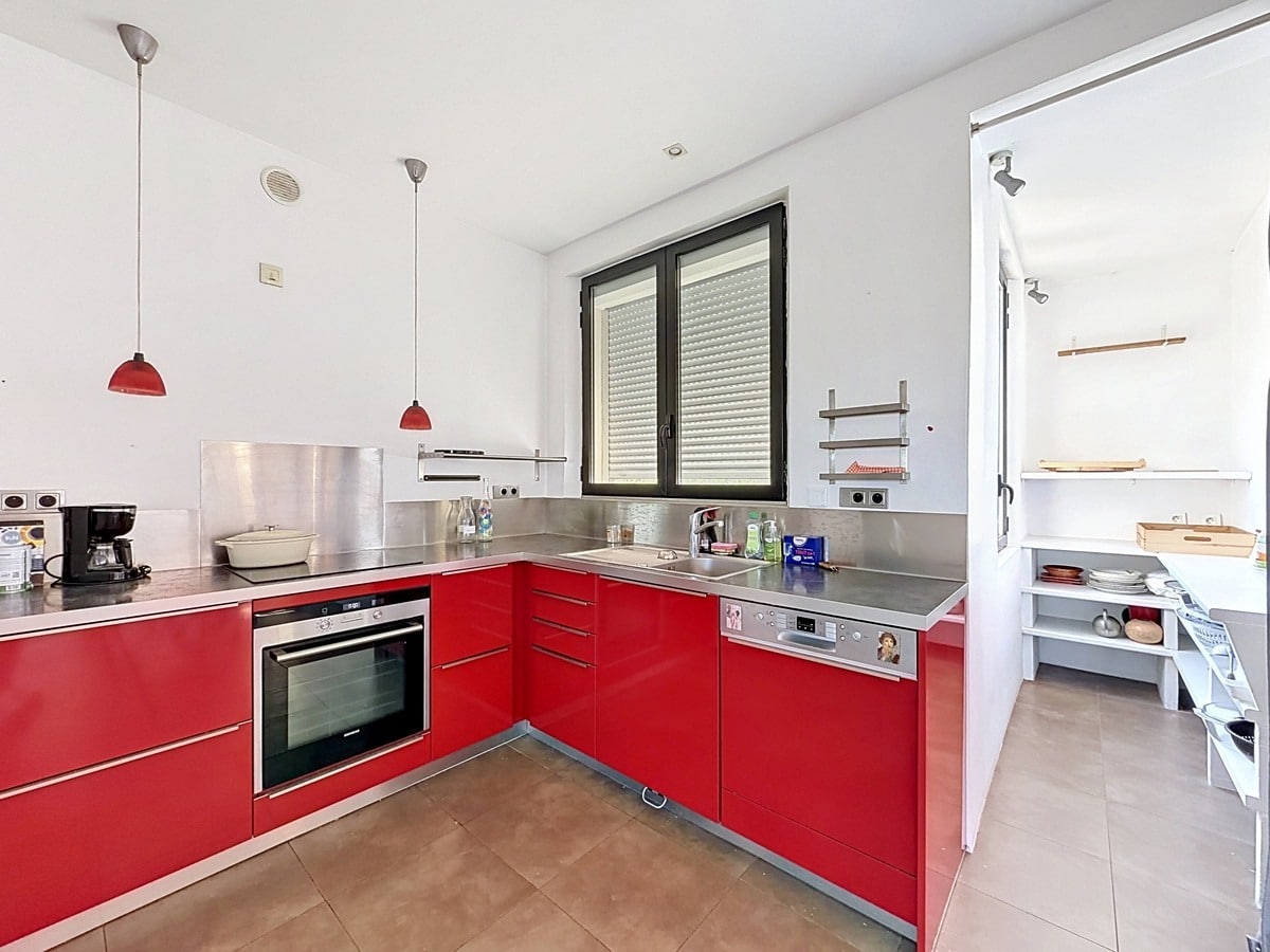 Cuisine en L avec meubles rouge brillant, plan de travail et crédence en inox brossé. Four encastré, lave-vaisselle intégré, suspensions rouges et cellier attenant visible à droite.