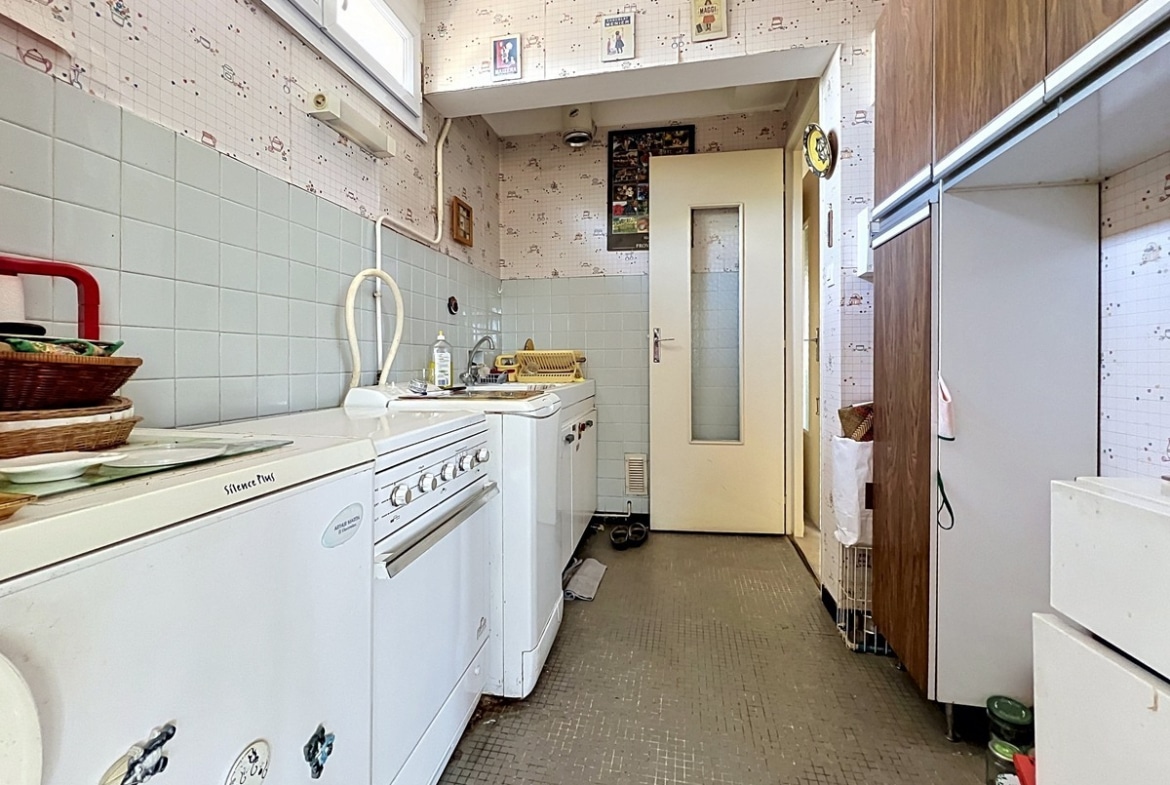 Cuisine en longueur avec carrelage mural vert pâle et papier peint à motifs. Cuisinière blanche, réfrigérateur, placards en bois foncé et sol en carrelage marron foncé mosaïque. Porte vitrée crème au fond menant vers un couloir.