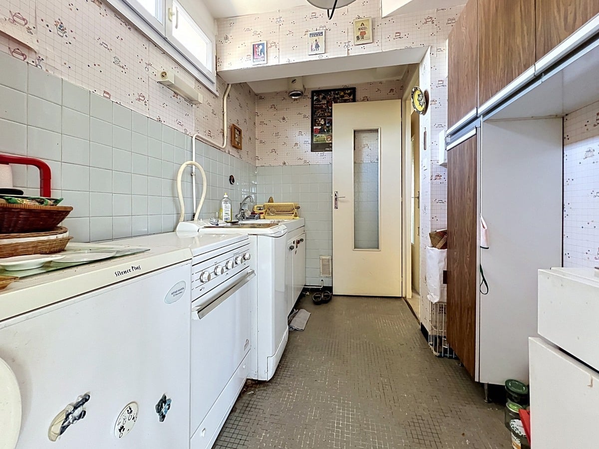 Cuisine en longueur avec carrelage mural vert pâle et papier peint à motifs. Cuisinière blanche, réfrigérateur, placards en bois foncé et sol en carrelage marron foncé mosaïque. Porte vitrée crème au fond menant vers un couloir.