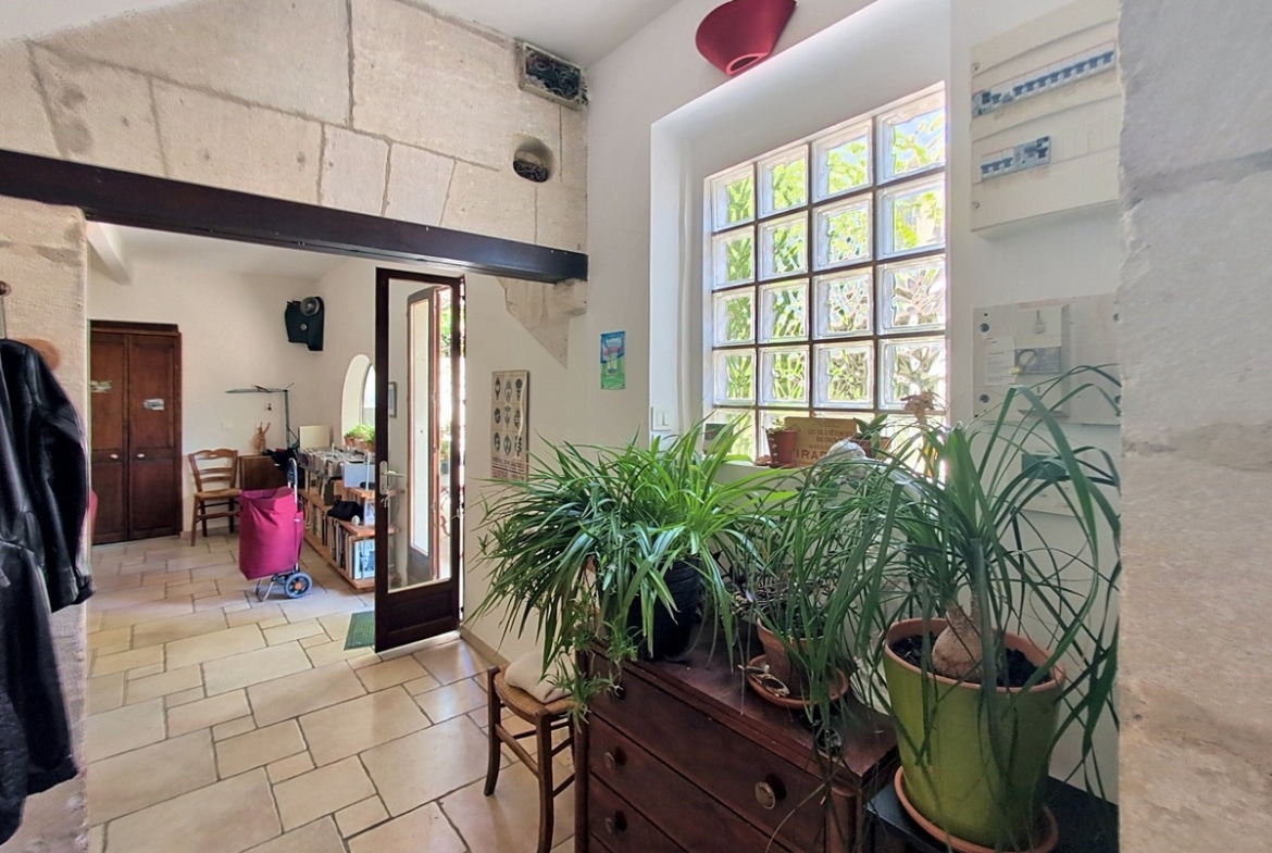 Hall d'entrée avec fenêtre en pavés de verre laissant filtrer la lumière naturelle, commode ancienne en bois supportant des plantes vertes. Sol en carrelage beige, murs en pierre et poutres métalliques apparentes.