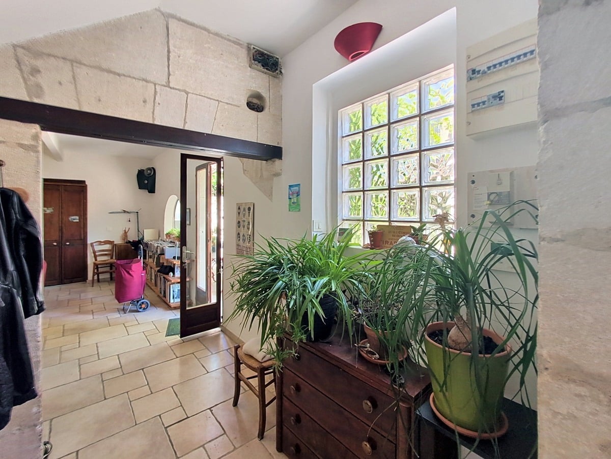 Hall d'entrée avec fenêtre en pavés de verre laissant filtrer la lumière naturelle, commode ancienne en bois supportant des plantes vertes. Sol en carrelage beige, murs en pierre et poutres métalliques apparentes.