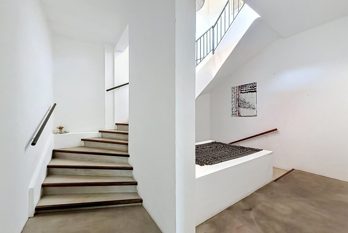 Cage d'escalier blanche avec marches en béton ciré et nez de marche en bois foncé. Mains courantes en bois, tableaux décoratifs aux murs, puits de lumière naturelle au plafond.