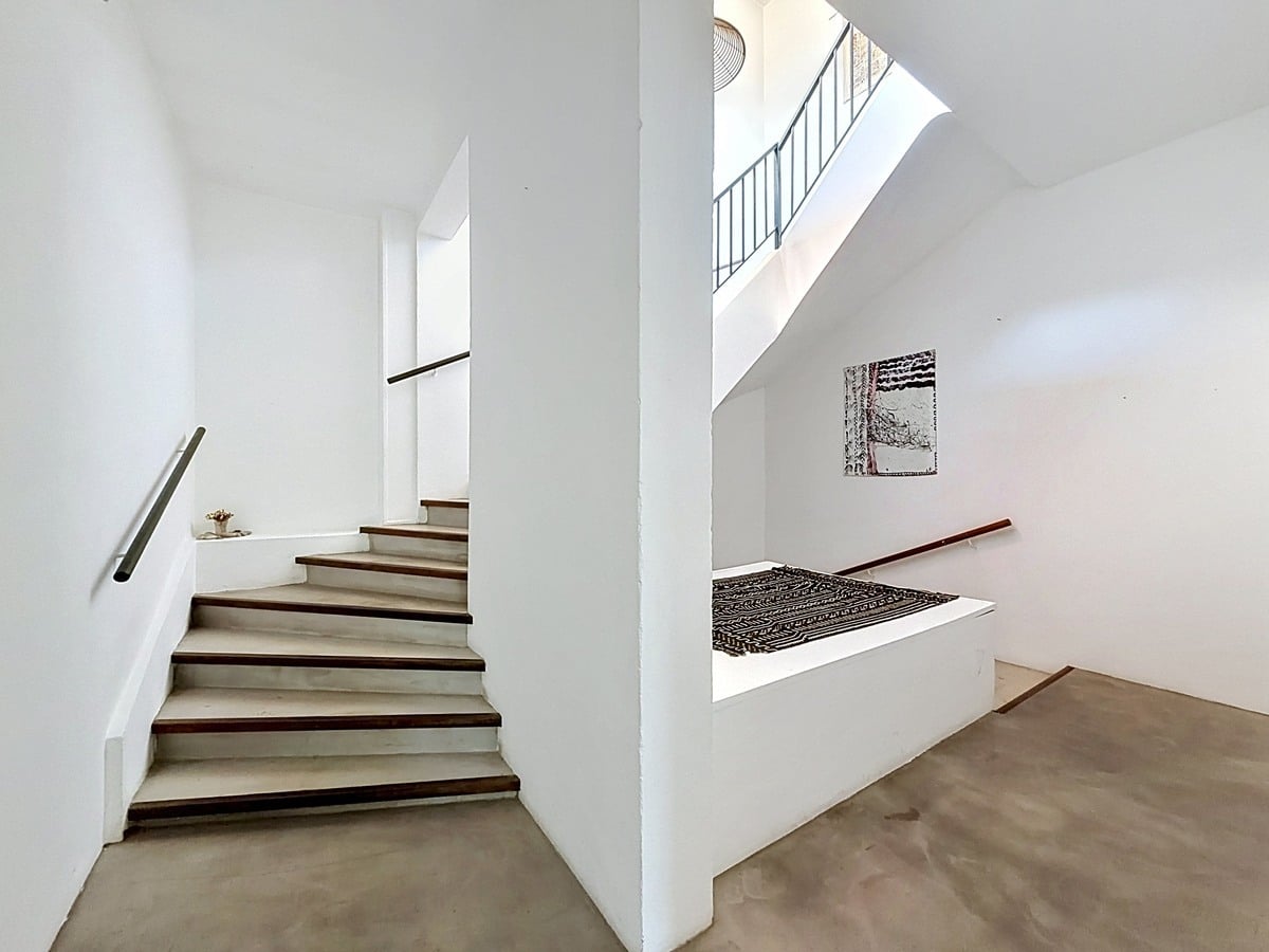 Cage d'escalier blanche avec marches en béton ciré et nez de marche en bois foncé. Mains courantes en bois, tableaux décoratifs aux murs, puits de lumière naturelle au plafond.