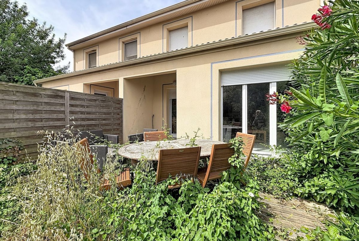 Façade arrière d'une maison à étage avec enduit ocre et volets roulants blancs. Baie vitrée coulissante au rez-de-chaussée donnant sur une terrasse couverte, jardin avec table en teck et végétation abondante incluant laurier rose fleuri et plantes grimpantes.