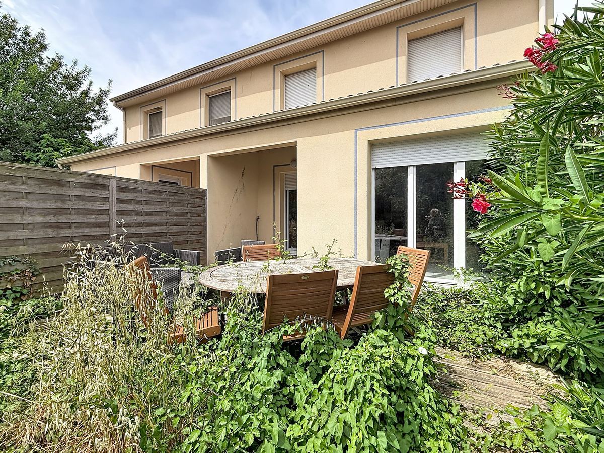 Façade arrière d'une maison à étage avec enduit ocre et volets roulants blancs. Baie vitrée coulissante au rez-de-chaussée donnant sur une terrasse couverte, jardin avec table en teck et végétation abondante incluant laurier rose fleuri et plantes grimpantes.
