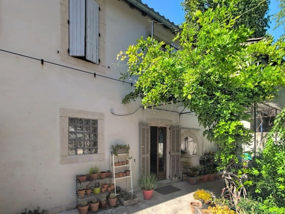 Façade extérieure en crépi beige avec volets gris-bleu, fenêtre en pavés de verre et porte d'entrée avec imposte en arche. Cour végétalisée avec plantes en pots, arbres feuillus et sol bétonné.
