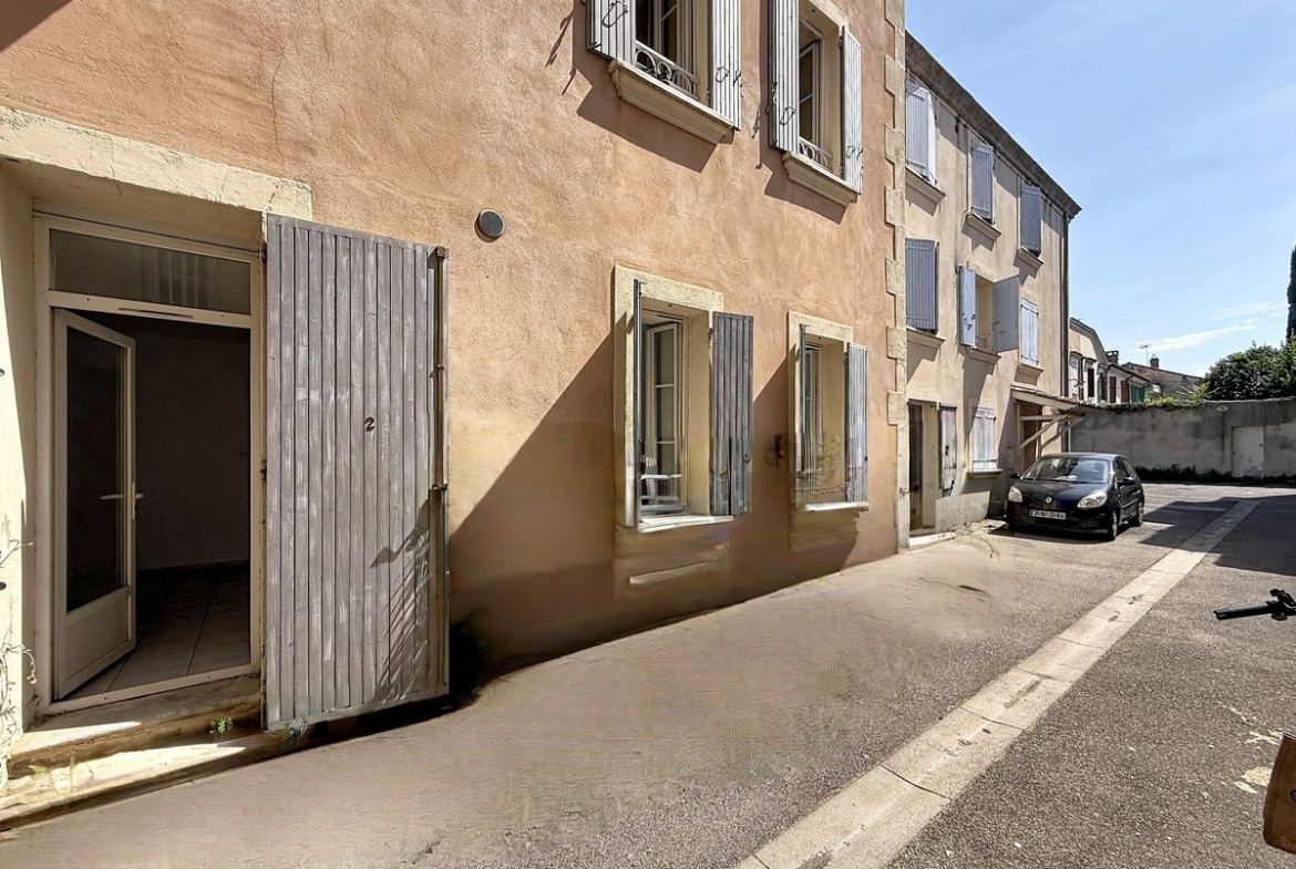 açade d'immeuble ancien en crépi ocre rose avec volets gris en bois sur une rue calme. L'entrée du rez-de-chaussée présente une porte vitrée et de grands volets battants gris ouverts.