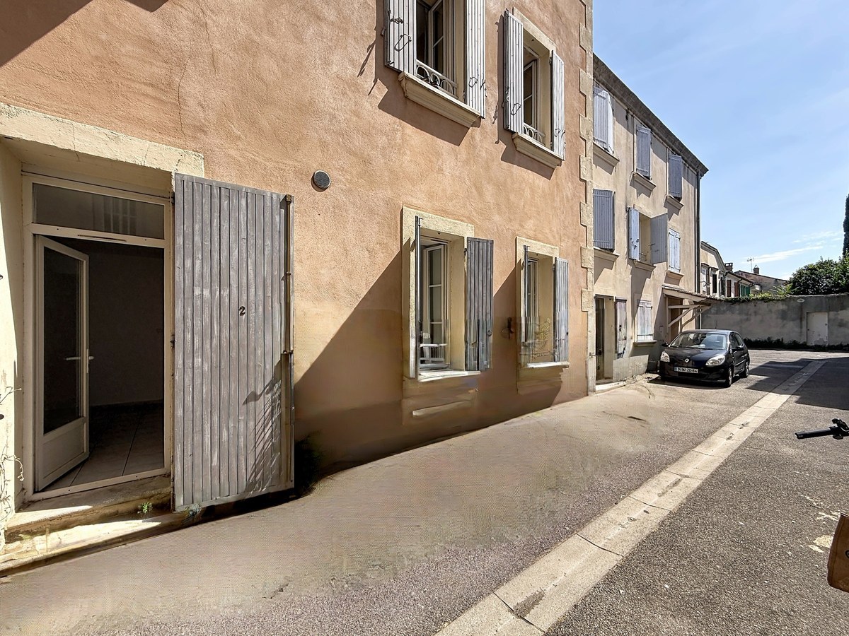 açade d'immeuble ancien en crépi ocre rose avec volets gris en bois sur une rue calme. L'entrée du rez-de-chaussée présente une porte vitrée et de grands volets battants gris ouverts.