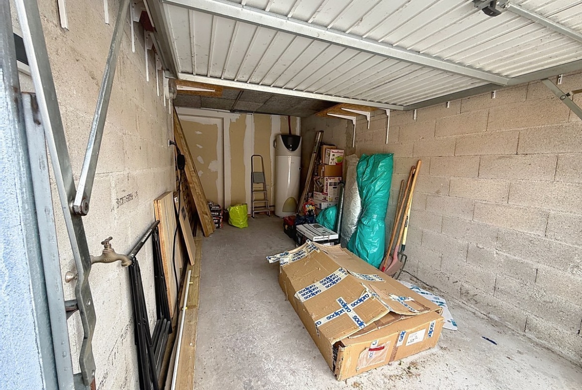 Garage en parpaings avec porte basculante métallique blanche et sol béton brut. Chauffe-eau vertical visible au fond, robinet extérieur sur le côté gauche, espace de stockage contenant escabeau, cartons et matériaux divers.
