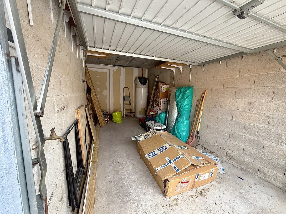 Garage en parpaings avec porte basculante métallique blanche et sol béton brut. Chauffe-eau vertical visible au fond, robinet extérieur sur le côté gauche, espace de stockage contenant escabeau, cartons et matériaux divers.